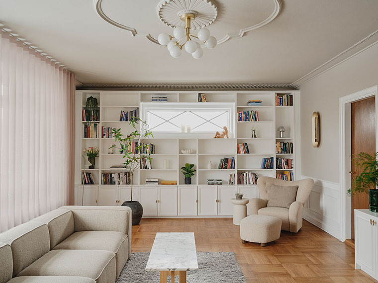 Lys stue med indbygget hvid reolvæg, beige sofa og lænestol, marmorbord og klassisk parketgulv.