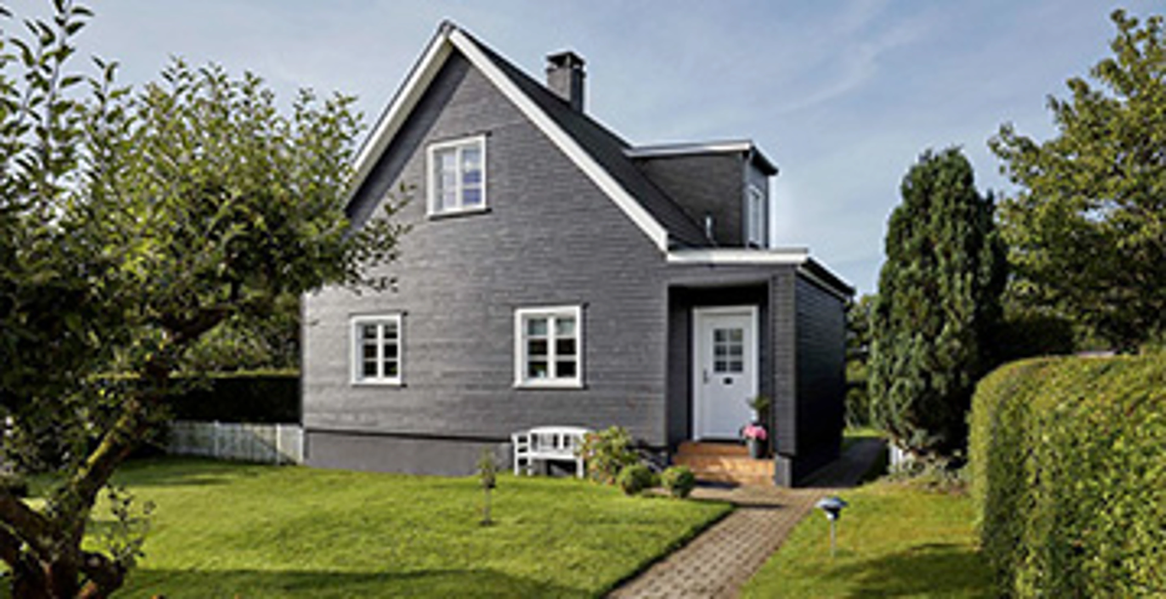 Villa med mörk fasad och vita fönsterkarmar, omgiven av en välskött trädgård. En stig leder genom gräsmattan fram till husets entré.