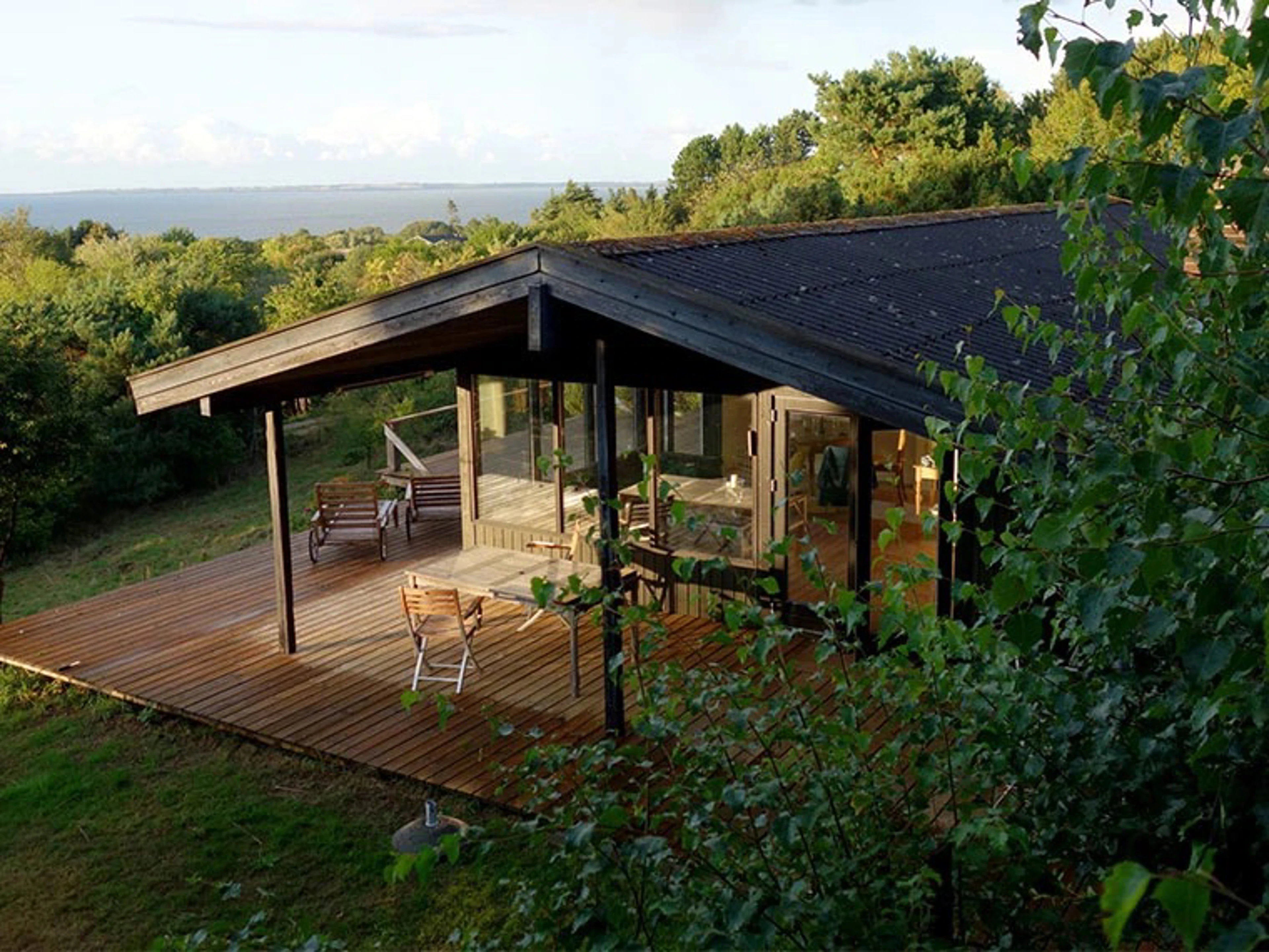 Moderne træhus med mørk facade, stor træterrasse og havudsigt omgivet af grøn natur.